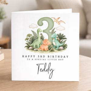 May include: A square white greeting card featuring a large green number 3, a green long-necked dinosaur, an orange triceratops, a green t-rex, and a flying pterodactyl. Text reads: HAPPY 3RD BIRTHDAY TO A SPECIAL LITTLE BOY and the name Teddy.