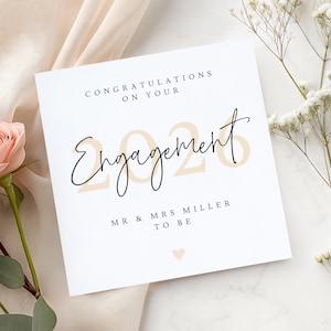 May include: A white engagement card with the words "Congratulations on Your Engagement" in black script. The year "2026" is in a light tan color. The card also includes "Mr & Mrs Miller To Be" and a small heart.