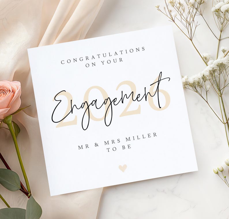 May include: A white engagement card with the words "Congratulations on Your Engagement" in black script. The year "2026" is in a light tan color. The card also includes "Mr & Mrs Miller To Be" and a small heart.