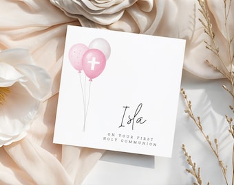 May include: A white card with the name "Isla" in cursive script, accompanied by the text "ON YOUR FIRST HOLY COMMUNION." The card features three watercolor balloons in pink and white, with a cross on one. The card is set against a soft, neutral background.