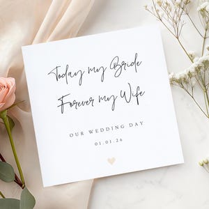 May include: A white square wedding card with black script that reads "Today my Bride, Forever my Wife." The card also includes "Our Wedding Day" and a date. A small peach heart is at the bottom. The card is surrounded by flowers.