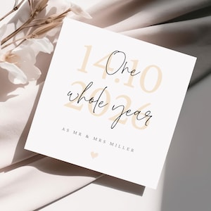 May include: A square white greeting card featuring the text One whole year in black script, overlaid on the large beige numbers 14.10.2020. Below this, the text reads AS MR & MRS MILLER with a small beige heart icon centred at the bottom.