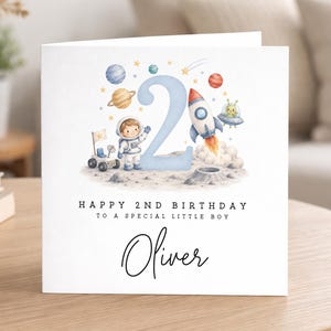 May include: A square greeting card featuring a watercolor space theme with a large light blue number 2, an astronaut, a rocket ship, planets, and a lunar rover. The text reads HAPPY 2ND BIRTHDAY TO A SPECIAL LITTLE BOY and the name Oliver.