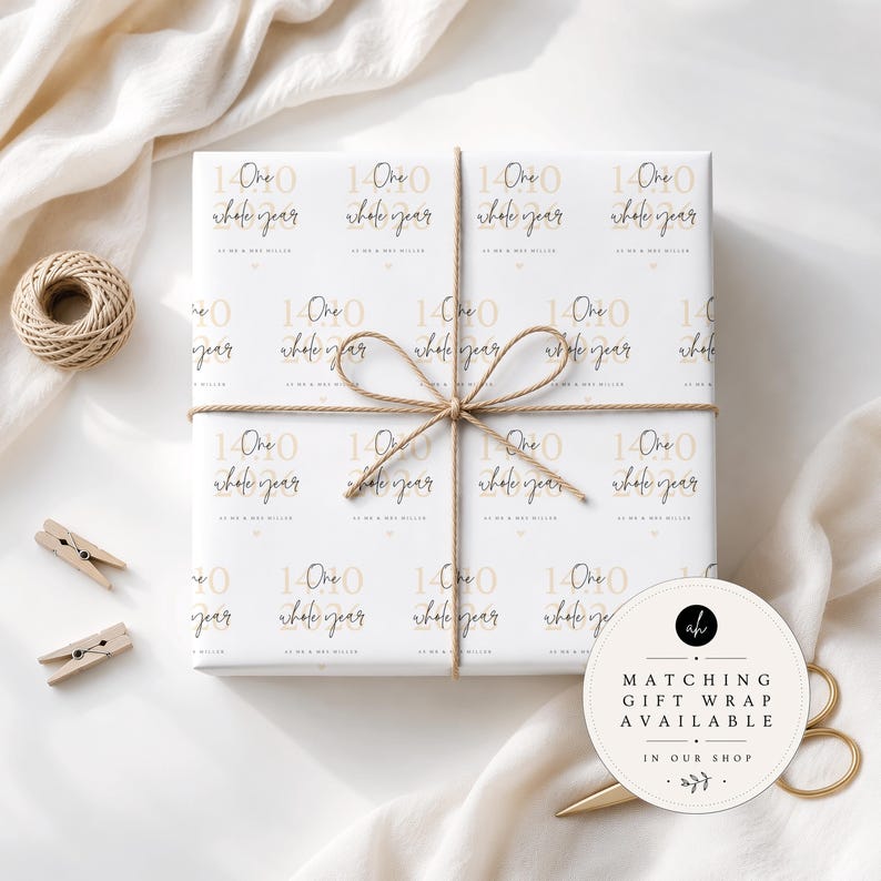 May include: A square gift box wrapped in white paper featuring a repeating pattern of the text 14.10.2020, One whole year, and As we & Mrs Miller in black and gold. The box is tied with thin brown twine in a bow centred on the top surface.