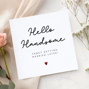 K&ouml;nnte beinhalten: Eine wei&szlig;e Gru&szlig;karte mit dem Text "Hello Handsome" in schwarzer Schrift. Darunter steht "Fancy getting married later?" mit einem kleinen roten Herz. Die Karte ist mit einer rosa Rose und wei&szlig;en Blumen verziert.
