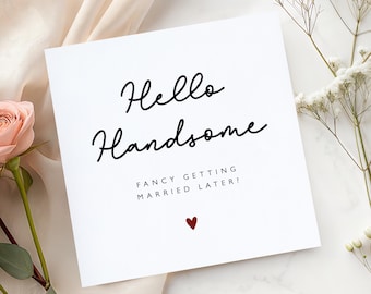 May include: A square white greeting card featuring the black cursive text Hello Handsome, followed by the capitalized black text FANCY GETTING MARRIED LATER? and a small red heart icon centered at the bottom of the card design.