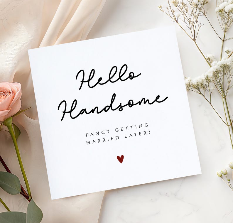 K&ouml;nnte beinhalten: Eine wei&szlig;e Gru&szlig;karte mit dem Text "Hello Handsome" in schwarzer Schrift. Darunter steht "Fancy getting married later?" mit einem kleinen roten Herz. Die Karte ist mit einer rosa Rose und wei&szlig;en Blumen verziert.
