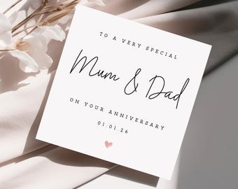 May include: A white square card with black script that reads "To a very special Mum & Dad on your anniversary 01.01.26" with a small pink heart at the bottom. The card is on a light beige background with white flowers.