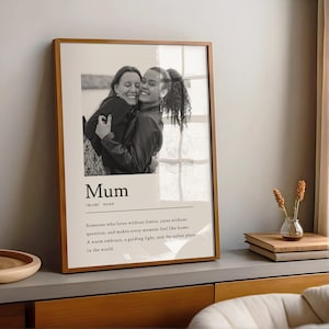 Op de afbeelding: Een zwart-witfoto in een bruine lijst van twee mensen die elkaar omhelzen. Onder de foto staat het woord "Mum" met een definitie. De afbeelding staat op een houten oppervlak.