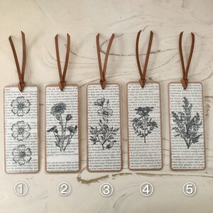 May include: Five rectangular bookmarks with brown leather cords. Each bookmark has a different black and white floral illustration on a background of vintage book pages. The edges of the bookmarks are brown.