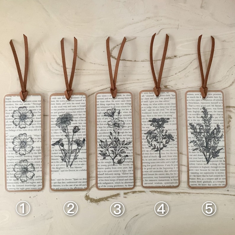 May include: Five rectangular bookmarks with brown leather cords. Each bookmark has a different black and white floral illustration on a background of vintage book pages. The edges of the bookmarks are brown.