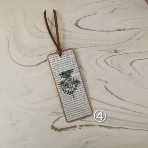 May include: A rectangular bookmark featuring a floral design and text from a book. It has a brown leather cord attached at the top. The floral design is black and white. The bookmark is made of paper.