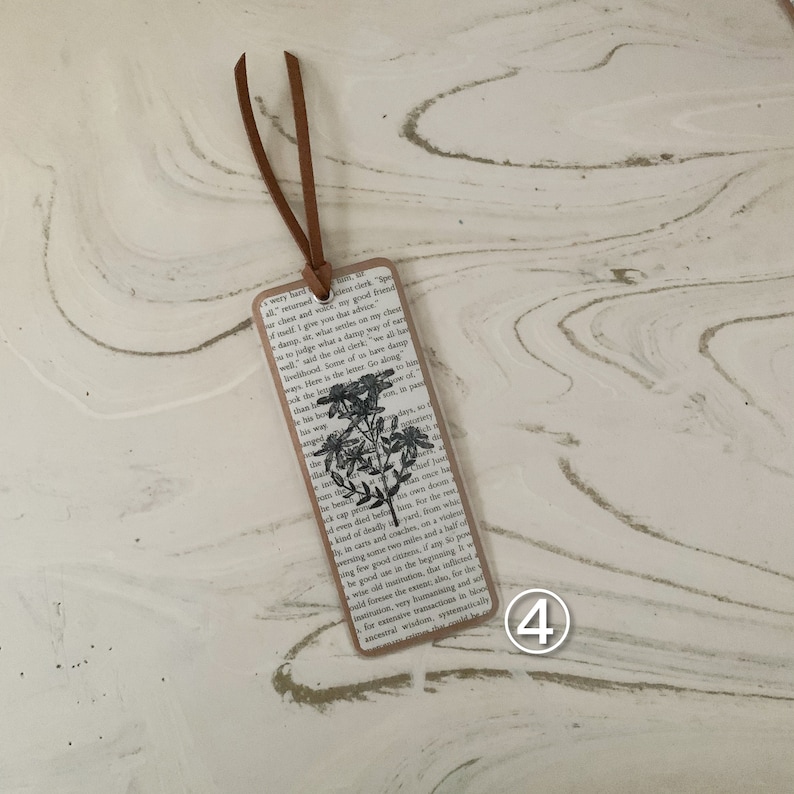 May include: A rectangular bookmark featuring a floral design and text from a book. It has a brown leather cord attached at the top. The floral design is black and white. The bookmark is made of paper.