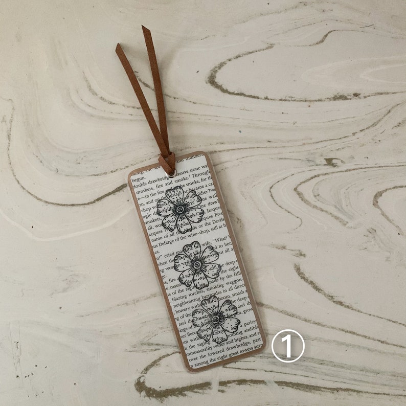 May include: A rectangular bookmark with a floral design, brown border, and leather cord. The background is a book page with visible text. Three black and white flower illustrations are printed on the bookmark. The bookmark measures approximately 15 cm in length.