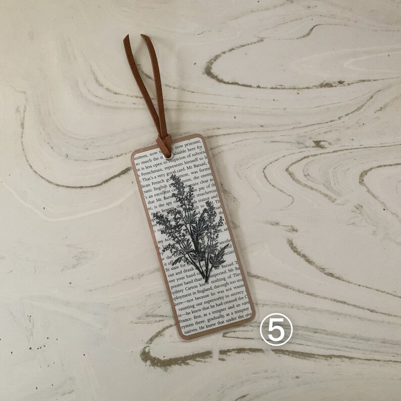 May include: A rectangular bookmark with a black and white botanical print. It has a brown leather cord and is made with a vintage book page background. The edges are trimmed with a brown border.