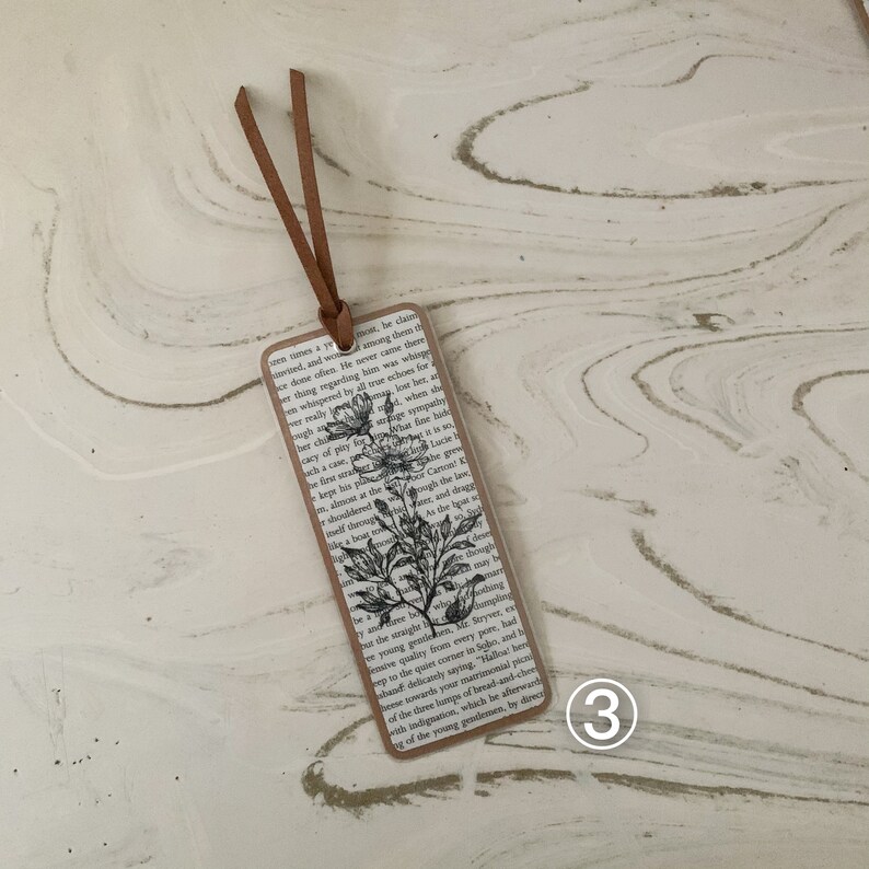 May include: A rectangular bookmark featuring a floral illustration and text from a book page. It has a brown border and a brown leather cord. The floral illustration is in black and white.