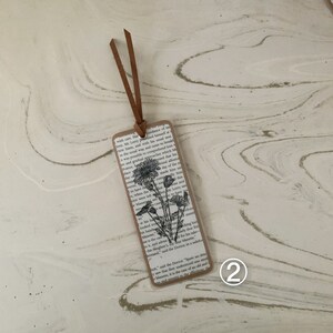 May include: A handcrafted bookmark with a floral design. The bookmark has a vintage appearance, with a printed page pattern and a brown leather cord. The floral illustration is a black and white drawing of daisies.