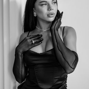 May include: A woman wearing a black satin dress with a plunging neckline and long black gloves. She is wearing a silver necklace and a ring on her finger. She has long dark hair and is looking up and to the right.