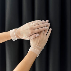 May include: A pair of white sheer gloves with delicate lace detailing. The gloves are worn on a person's hands, with the fingers extended and the hands clasped together.