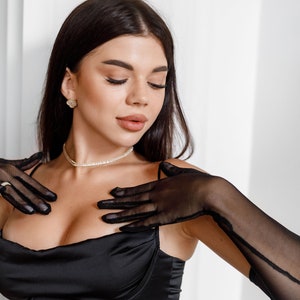 May include: A woman wearing a black satin top with a low neckline and long black sheer gloves. She is wearing a pearl necklace and small gold earrings.