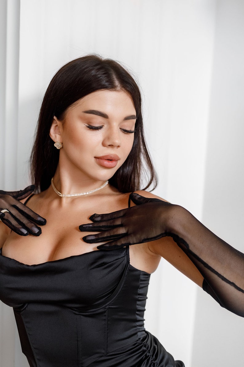 May include: A woman wearing a black satin top with a low neckline and long black sheer gloves. She is wearing a pearl necklace and small gold earrings.
