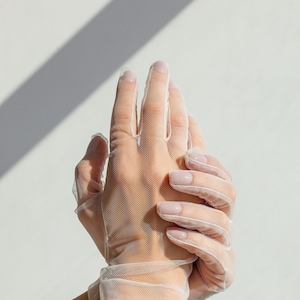 May include: A pair of white sheer gloves with delicate netting. The gloves are worn on a person's hands, with the fingers extended and the hands clasped together.