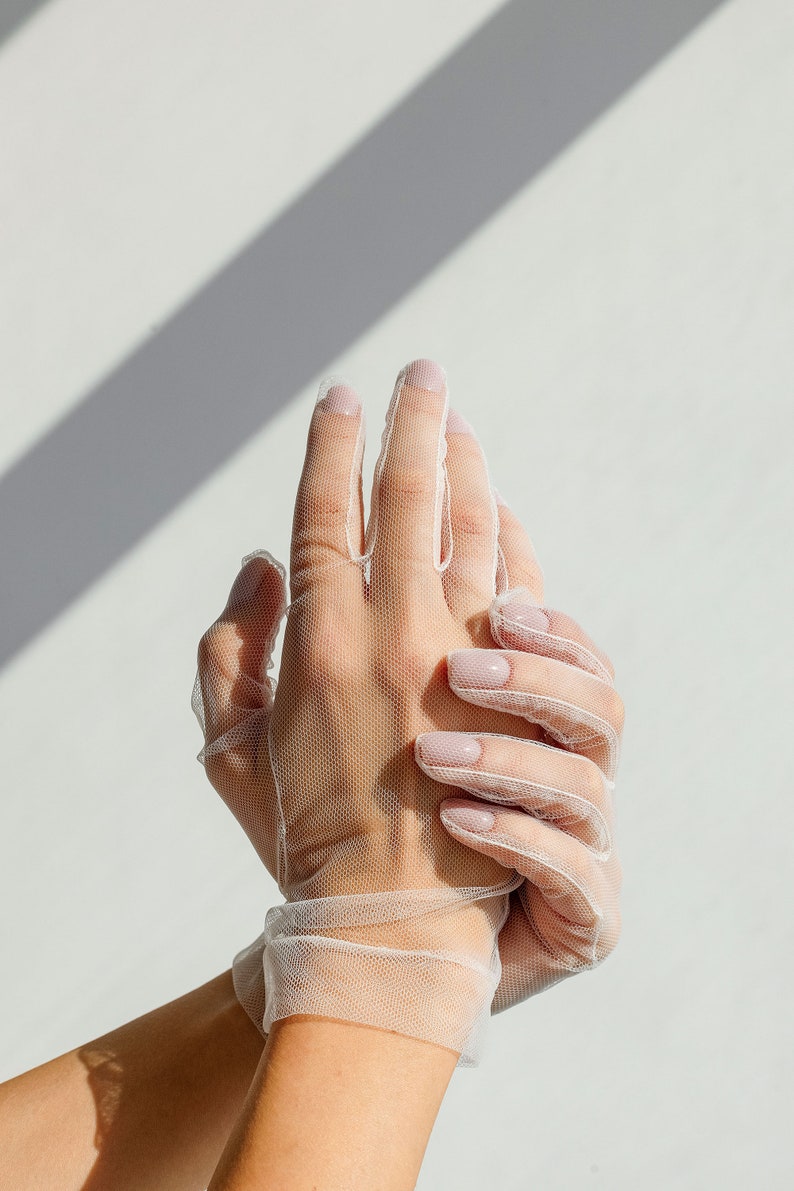 May include: A pair of white sheer gloves with delicate netting. The gloves are worn on a person's hands, with the fingers extended and the hands clasped together.