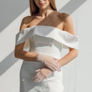 May include: A white off-the-shoulder wedding dress with a fitted bodice and a flowing skirt. The dress has a simple design and is accented with delicate lace gloves.