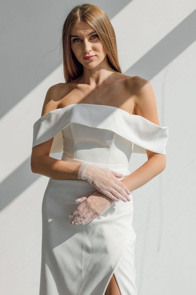 May include: A white off-the-shoulder wedding dress with a fitted bodice and a flowing skirt. The dress has a simple design and is accented with delicate lace gloves.