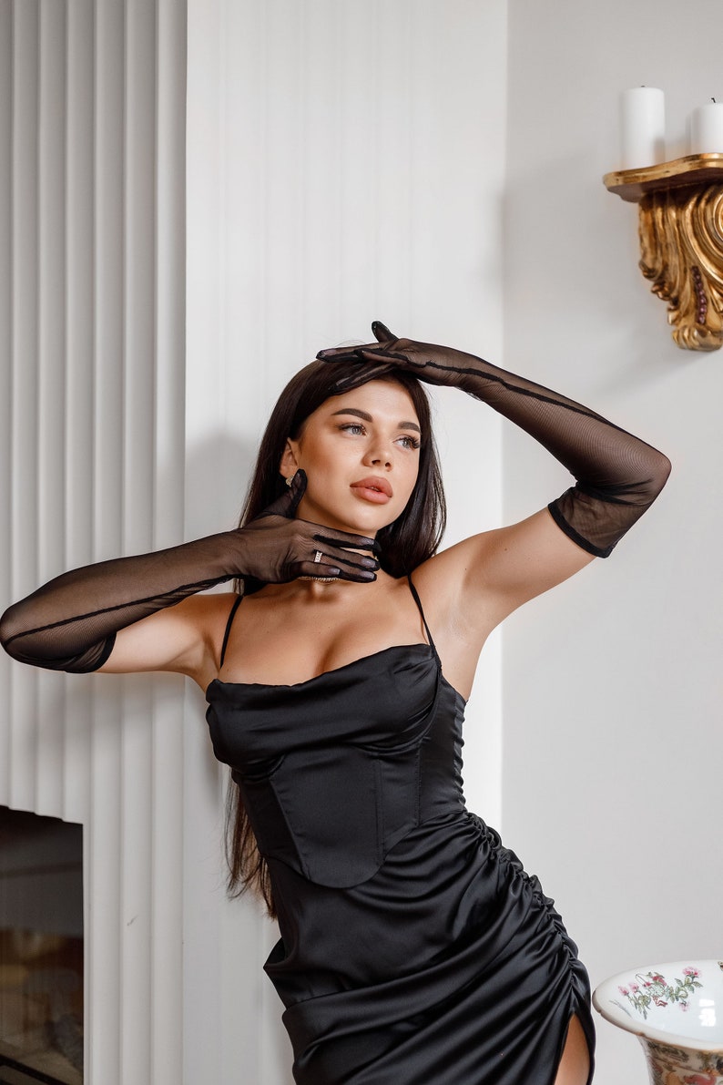 May include: A woman wearing a black satin dress with a cowl neck and a pair of black sheer gloves. She is standing in front of a white wall with a gold decorative element on the wall.