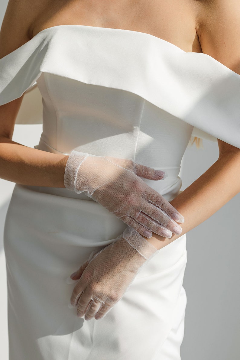 May include: A white off-the-shoulder dress with a pair of white sheer gloves.