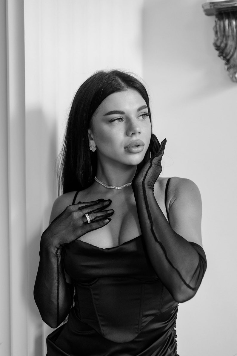 May include: A woman wearing a black satin dress with a plunging neckline and long black gloves. She is wearing a silver necklace and a ring on her finger. She has long dark hair and is looking up and to the right.