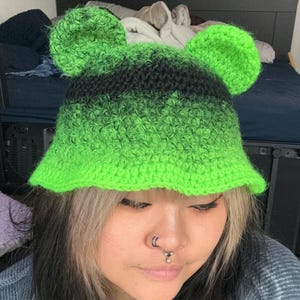 May include: A handmade, green and black crochet hat with bear ears. The hat features a gradient design, transitioning from bright green to black. The hat is a bucket style, with a rounded crown and a slightly flared brim.