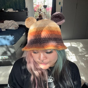 May include: A handmade crochet hat with a bear ear design. The hat features a color gradient from light tan to dark brown, with two large, round ears on top. The brim is slightly flared. The hat is a unique, cozy accessory.