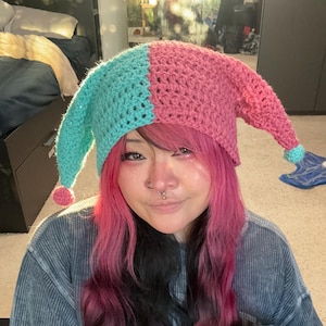 May include: A two-toned crochet jester hat featuring a split design with one side in bright turquoise and the other in dusty rose pink. The hat has two long, pointed ear-like flaps extending from the sides, each finished with a small pom-pom.