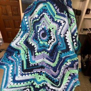 Op de afbeelding: Een handgemaakte, stervormige gehaakte deken in tinten blauw, groen en paars. De deken heeft een gedetailleerd, open ontwerp met een geschulpte rand. Het kleurenpalet omvat teal, marineblauw en lichtgroen, waardoor een visueel aantrekkelijk textiel ontstaat.