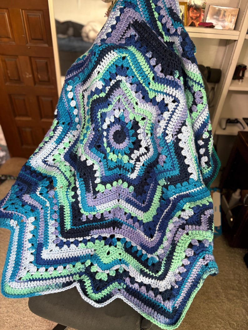 Op de afbeelding: Een handgemaakte, stervormige gehaakte deken in tinten blauw, groen en paars. De deken heeft een gedetailleerd, open ontwerp met een geschulpte rand. Het kleurenpalet omvat teal, marineblauw en lichtgroen, waardoor een visueel aantrekkelijk textiel ontstaat.