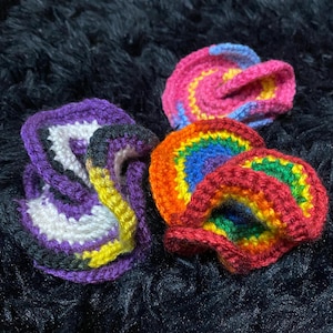 May include: Close-up of three colourful crocheted items. One is purple, white, yellow, and black. Another is pink, blue, and yellow. The third is a rainbow of colours, including red, orange, yellow, green, and blue.