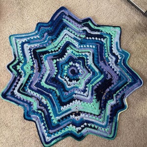 Op de afbeelding: Een handgemaakte, stervormige gehaakte deken in tinten blauw, turquoise en paars. De deken heeft een gedetailleerd, gelaagd ontwerp met een geschulpte rand. Het garen lijkt zacht en de deken wordt waarschijnlijk gebruikt voor decoratieve of functionele doeleinden.
