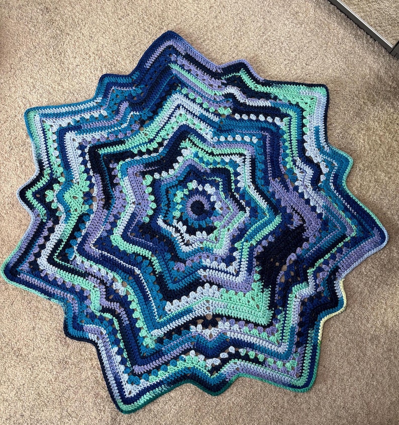 Op de afbeelding: Een handgemaakte, stervormige gehaakte deken in tinten blauw, turquoise en paars. De deken heeft een gedetailleerd, gelaagd ontwerp met een geschulpte rand. Het garen lijkt zacht en de deken wordt waarschijnlijk gebruikt voor decoratieve of functionele doeleinden.