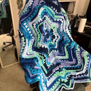 Op de afbeelding: Een handgemaakte, stervormige gehaakte deken in tinten blauw, teal en lavendel. De deken heeft een gedetailleerd geometrisch patroon met een geschulpte rand. De deken is over een stoel gedrapeerd.