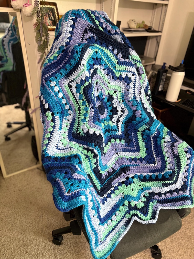 Op de afbeelding: Een handgemaakte, stervormige gehaakte deken in tinten blauw, teal en lavendel. De deken heeft een gedetailleerd geometrisch patroon met een geschulpte rand. De deken is over een stoel gedrapeerd.