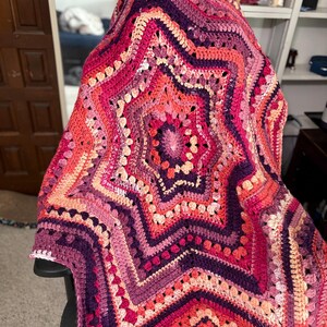 以下が含まれることがあります： ピンク、紫、ピーチの色合いの手作りの星型の編み物ブランケット。ブランケットは、複数の層と中心のスターバーストパターンを備えた、詳細でテクスチャ加工されたデザインが特徴です。ブランケットは椅子に掛けられています。