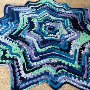 Op de afbeelding: Een handgemaakte, stervormige gehaakte deken in tinten blauw, turquoise en paars. De deken heeft een gedetailleerd, gelaagd ontwerp met een geschulpte rand. Het garen lijkt zacht, met een diameter van ongeveer 120 cm.