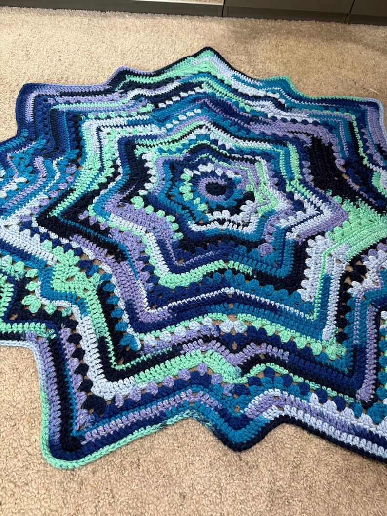 Op de afbeelding: Een handgemaakte, stervormige gehaakte deken in tinten blauw, turquoise en paars. De deken heeft een gedetailleerd, gelaagd ontwerp met een geschulpte rand. Het garen lijkt zacht, met een diameter van ongeveer 120 cm.