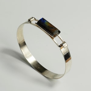 May include: A silver bangle bracelet featuring a rectangular, multi-colored stone. The stone displays hues of blue, brown, and yellow. The bracelet has a simple, elegant design with a clasp closure. The metal has a polished finish.