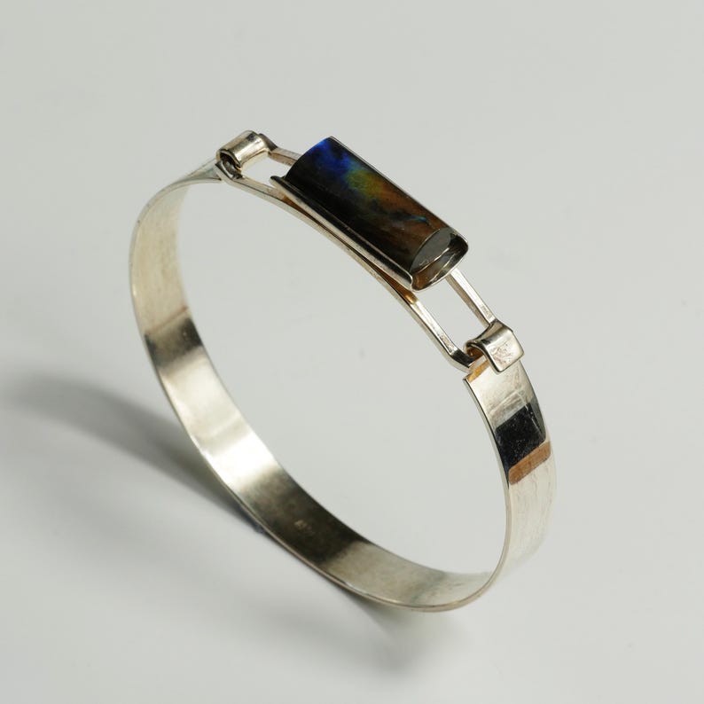 May include: A silver bangle bracelet featuring a rectangular, multi-colored stone. The stone displays hues of blue, brown, and yellow. The bracelet has a simple, elegant design with a clasp closure. The metal has a polished finish.