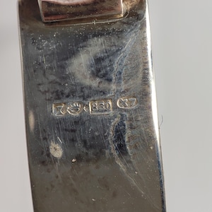 May include: Close-up of a silver metal object with visible hallmarks. The hallmarks include the number "830" and other symbols. The metal has a polished, reflective surface with some signs of wear.