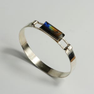 May include: A silver bracelet with a rectangular, iridescent stone at the center. The stone displays a mix of blue, yellow, and brown hues. The bracelet has a simple, elegant design with a clasp closure. A piece of jewelry.
