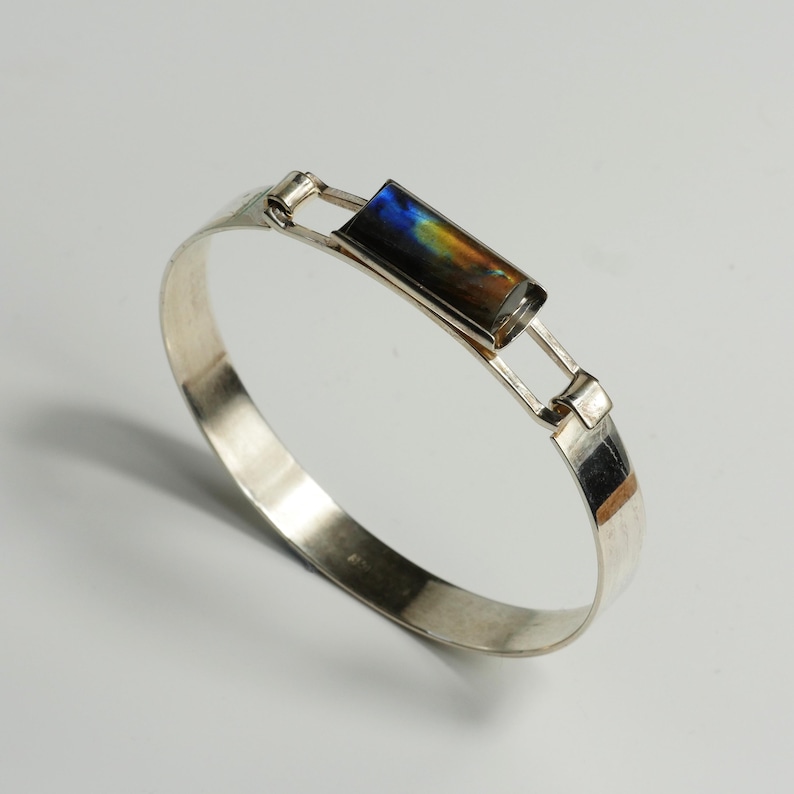 May include: A silver bracelet with a rectangular, iridescent stone at the center. The stone displays a mix of blue, yellow, and brown hues. The bracelet has a simple, elegant design with a clasp closure. A piece of jewelry.